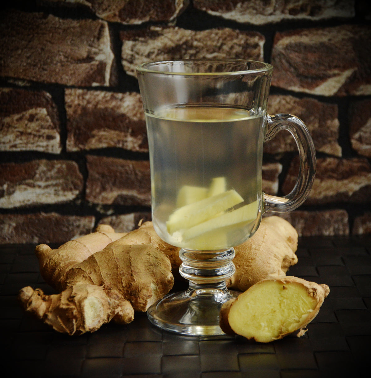 A clear glass mug of ginger tea with slices of ginger inside, beside whole fresh ginger root in front of a stone wall