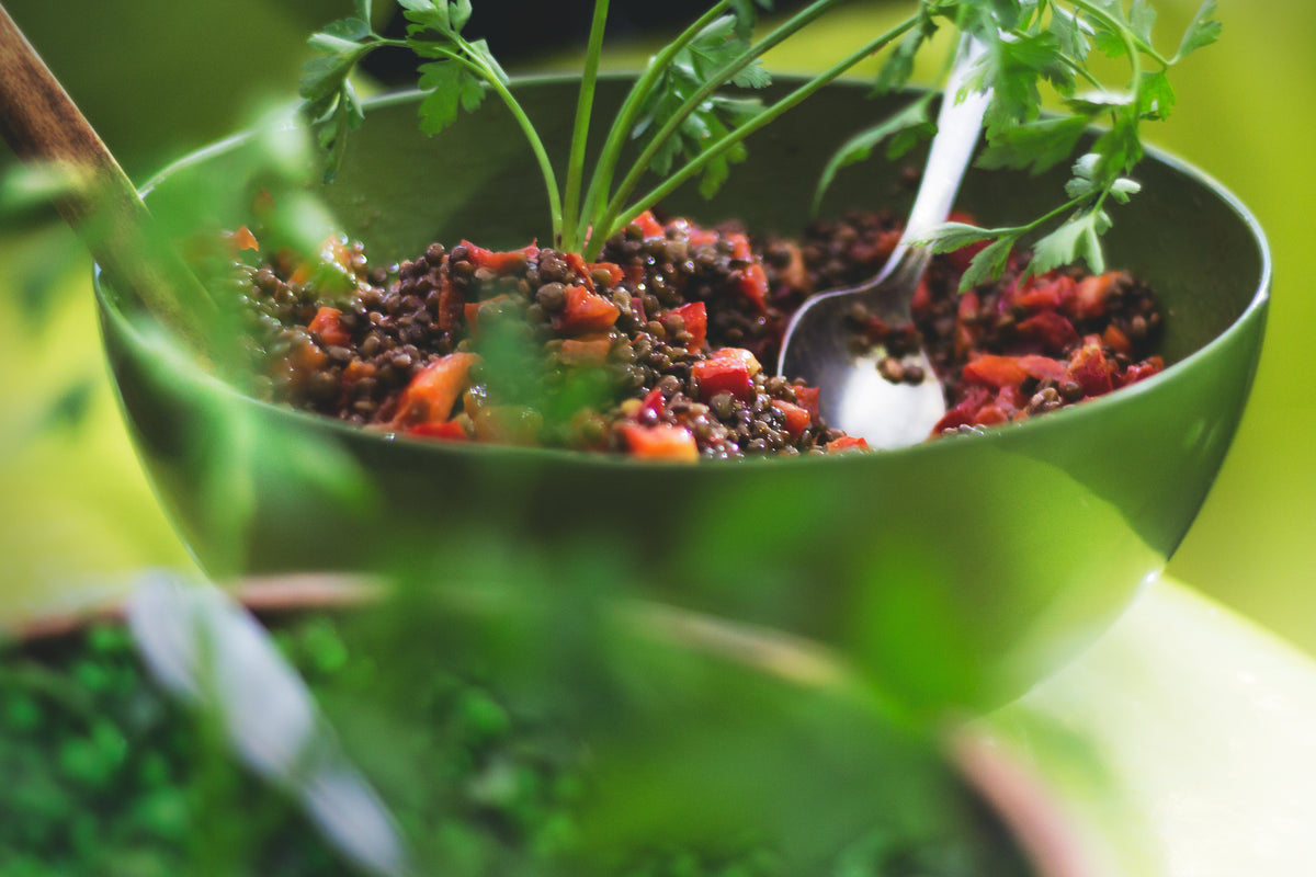 A green bowl of dark lentil salad with diced red peppers and fresh parsley, a metal spoon resting in it
