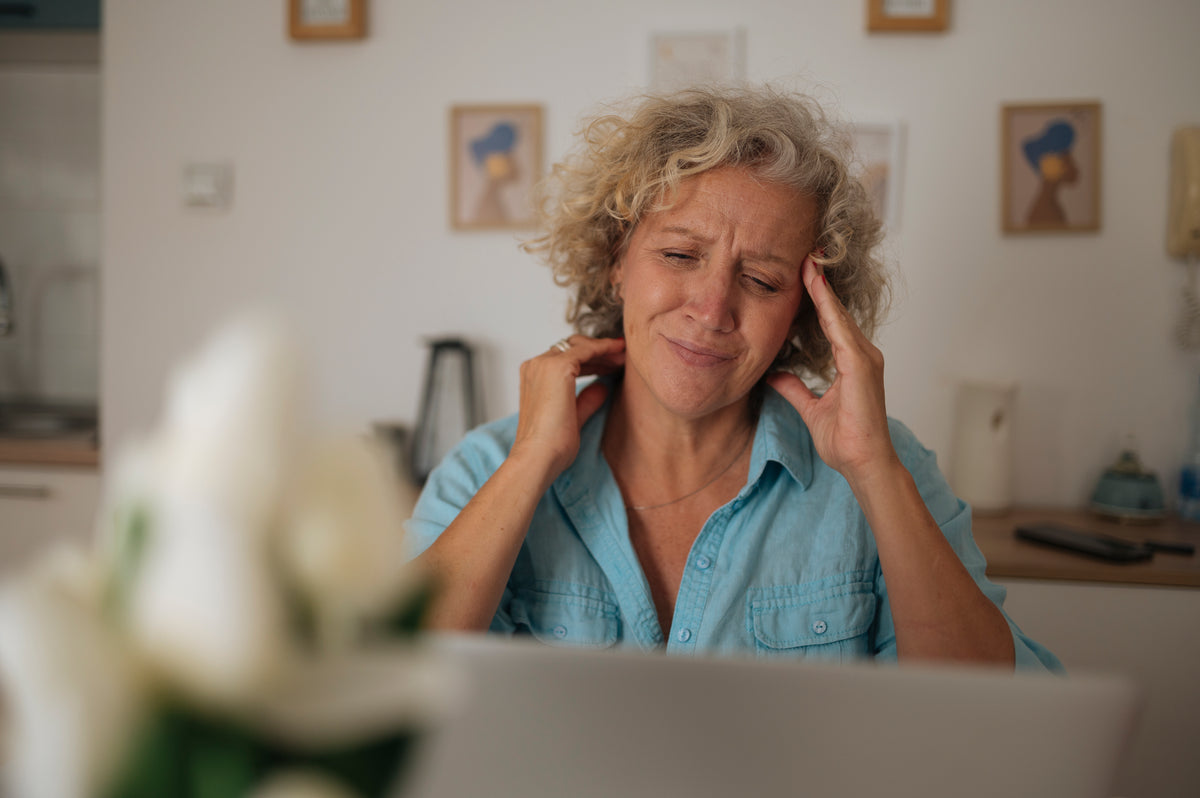 A woman with curly gray-blonde hair in a light blue shirt winces at her laptop, one hand pressed to her temple and the other on her neck