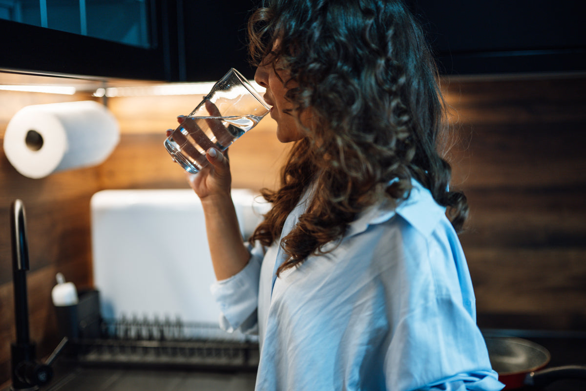 A woman with curly hair drinks a glass of water in a kitchen