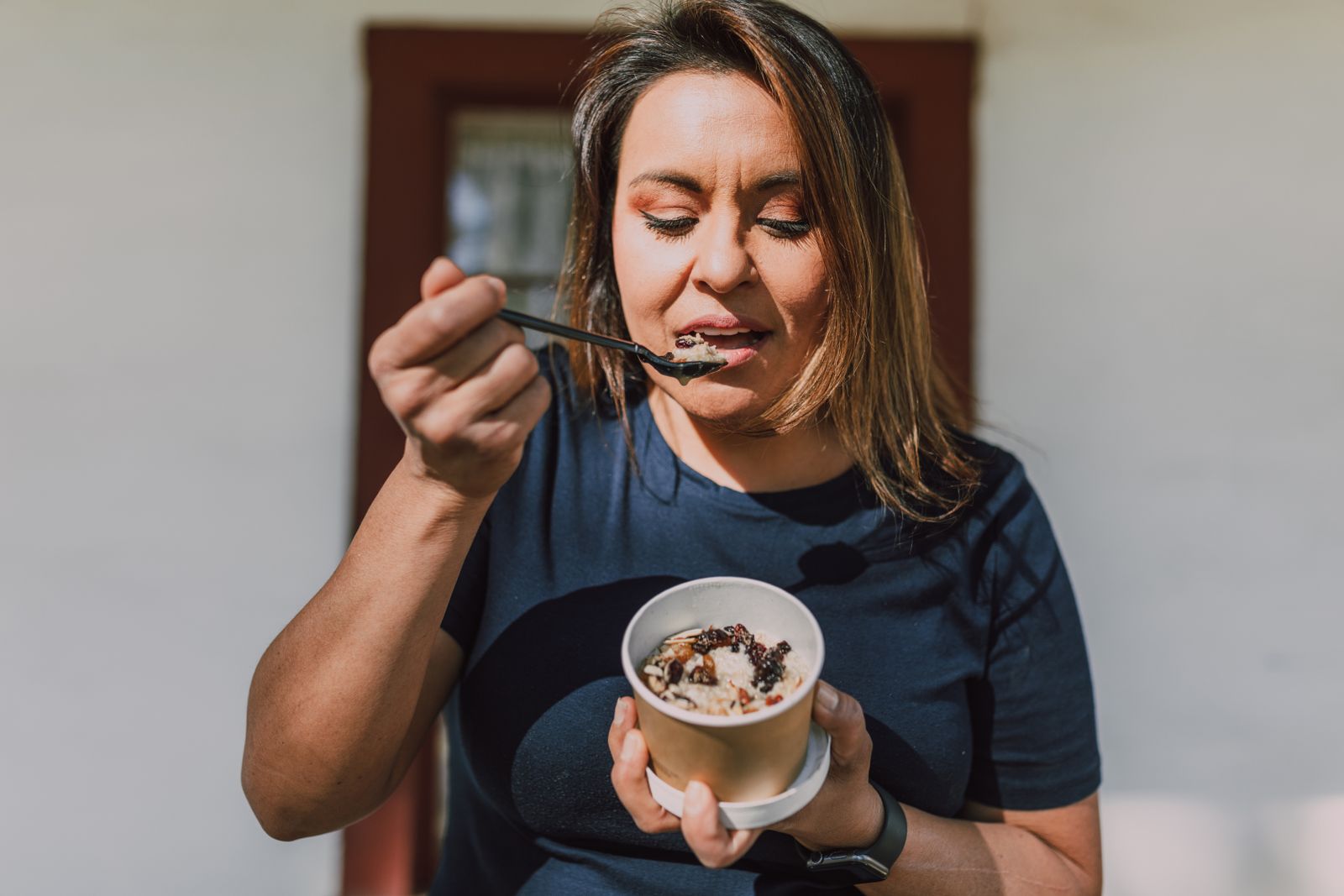 Woman holding and eating a bowl of oatmeal topped with fruit in daylight, a high-fiber breakfast